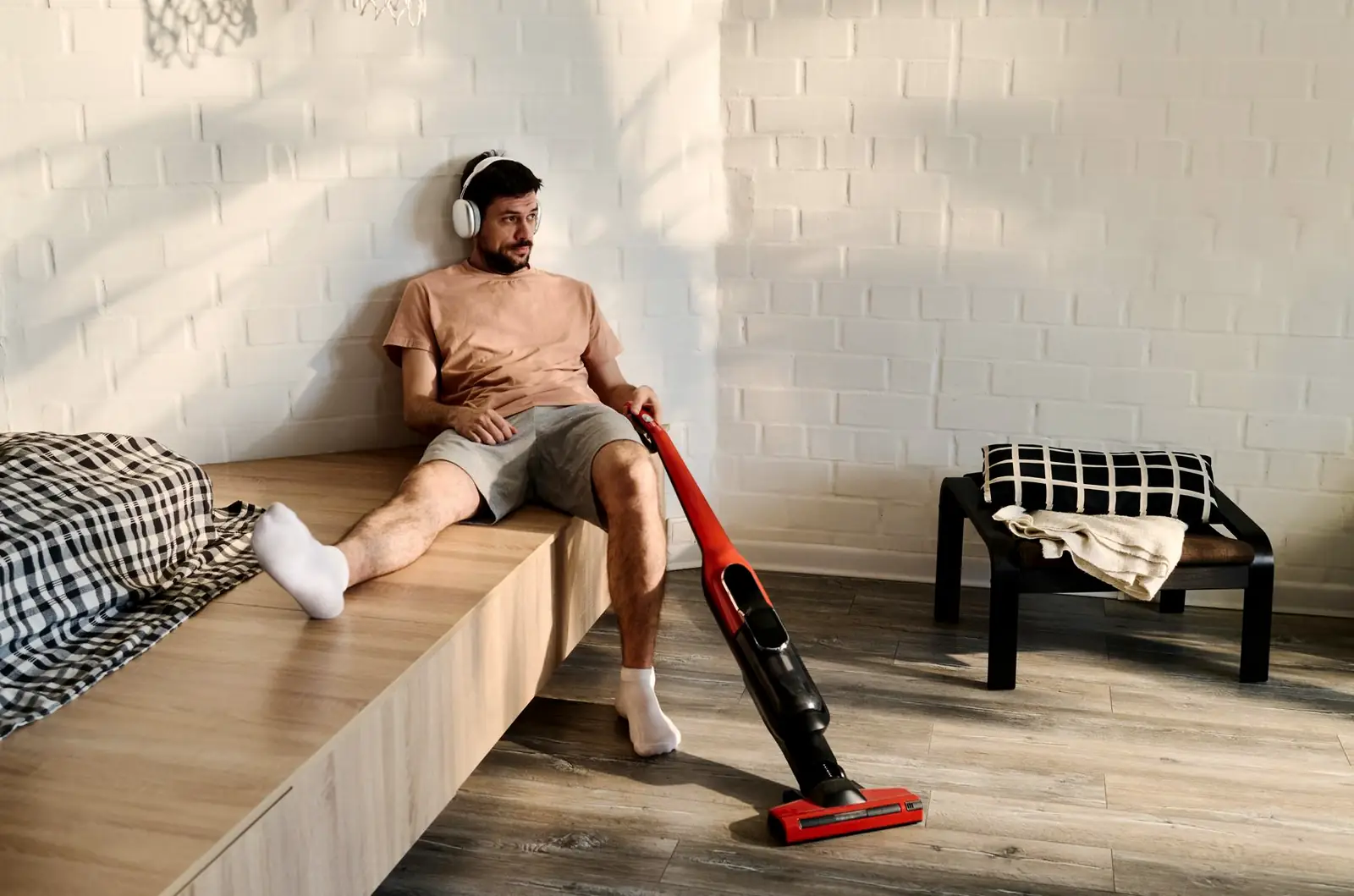 A person sitting on a wooden bench-bed next to a cordless vacuum cleaner in a plain bedroom with checkered bedding