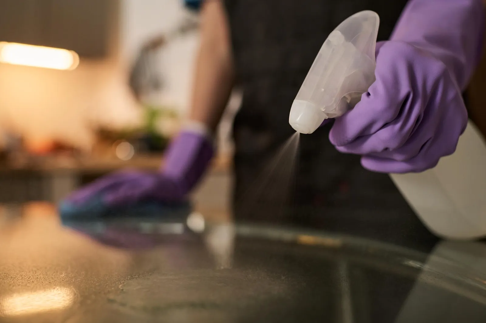 A purple-gloved hand spraying cleaner over a dark worktop while the other hand wipes with a cloth